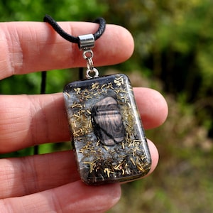 Puede incluir: Un colgante rectangular con un cordón negro. El colgante es de resina transparente con una piedra oscura e inclusiones doradas. La piedra tiene forma ovalada con patrones marrones oscuros y claros. El colgante se sujeta con una fianza plateada.