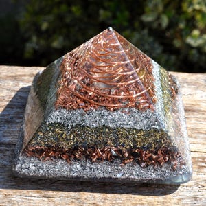 Peut inclure: Une pyramide en résine transparente avec des couches de matériaux argentés, dorés et cuivrés. Le sommet présente une spirale en cuivre. La pyramide est posée sur une surface en bois. C'est un objet décoratif.