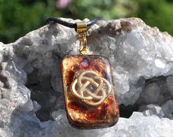 May include: A rectangular pendant featuring a gold-colored Celtic knot emblem embedded in a translucent amber and brown resin base with gold flecks, suspended from a black cord by a gold-toned metal bail.