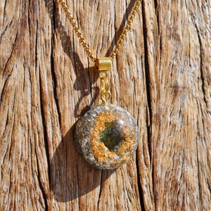 Op de afbeelding: Een ronde hanger aan een goudkleurige ketting met een harsbedel. De bedel bevat ingebedde metallic goudvlokken en donkergroene mineraalfragmenten, opgehangen aan een goudkleurig metalen oogje dat aan de ketting is bevestigd.