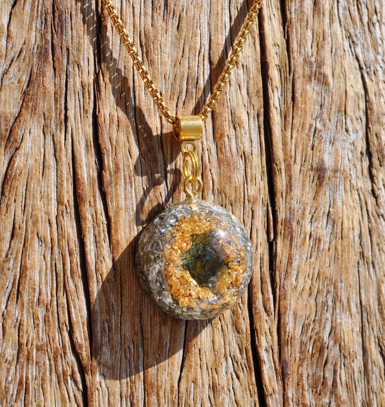 Op de afbeelding: Een ronde hanger aan een goudkleurige ketting met een harsbedel. De bedel bevat ingebedde metallic goudvlokken en donkergroene mineraalfragmenten, opgehangen aan een goudkleurig metalen oogje dat aan de ketting is bevestigd.