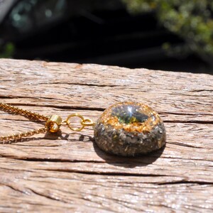 Op de afbeelding: Een goudkleurige box-ketting met een koepelvormige hanger. De hanger heeft een donkere, getextureerde basis met een ring van goudkleurige vlokken rondom een glad, doorschijnend, olijfgroen middendeel dat fel zonlicht reflecteert.