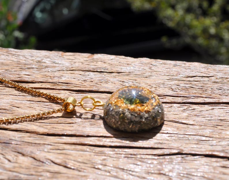 Op de afbeelding: Een goudkleurige box-ketting met een koepelvormige hanger. De hanger heeft een donkere, getextureerde basis met een ring van goudkleurige vlokken rondom een glad, doorschijnend, olijfgroen middendeel dat fel zonlicht reflecteert.
