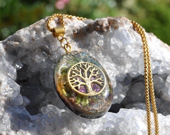 May include: An oval-shaped pendant featuring an embedded gold-toned tree of life emblem, suspended from a gold-toned box chain. The pendant contains a mix of green, purple, and metallic flecks within a clear resin base, creating a layered appearance.