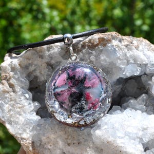 Könnte beinhalten: Eine transparente, runde Anhängerkette mit einem schwarzen Band. Der Anhänger enthält rosa, schwarze und silberne Elemente. Die Halskette wird vor einer weißen und grauen Felsformation mit verschwommenem grünem Hintergrund präsentiert.