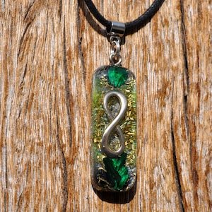 May include: A rectangular pendant with a silver infinity symbol encased in a clear resin. The resin contains green and gold flecks, with green accents at the top and bottom. The pendant hangs from a black cord.