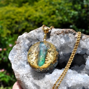 Può includere: Un ciondolo circolare con un frammento di cristallo verde iridescente al centro, incastonato in una base di resina scura con scaglie di foglia d'oro. Il ciondolo è fissato a una catena a corda dorata e poggia su una grande geode di cristallo.