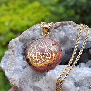 May include: A round pendant featuring a gold-colored lotus-like geometric design embedded in a reddish-brown resin base. The pendant is attached to a gold and silver-toned twisted rope chain, creating a metallic contrast in the jewelry piece.