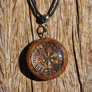Op de afbeelding: Een ronde hanger met een zilverkleurig Vegvisir-symbool ingebed in een doorschijnende, amberkleurige harsbasis gevuld met gouden glitters. De hanger hangt aan een zwart koord met een zilverkleurig metalen oogje aan de bovenzijde.