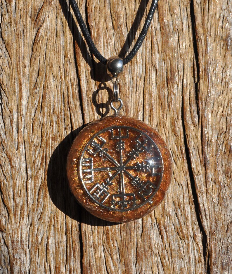 Op de afbeelding: Een ronde hanger met een zilverkleurig Vegvisir-symbool ingebed in een doorschijnende, amberkleurige harsbasis gevuld met gouden glitters. De hanger hangt aan een zwart koord met een zilverkleurig metalen oogje aan de bovenzijde.