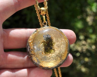 May include: A circular pendant featuring a clear resin center with embedded gold leaf flakes and a dark green mineral fragment, encased in a textured metallic border. The pendant hangs from a gold-toned twisted rope chain necklace.