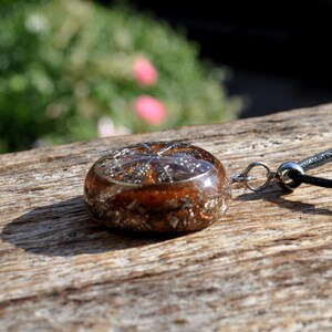 Op de afbeelding: Een ronde hanger met een heldere harskoepel gevuld met koperkleurige metaalvlokken en een zilverkleurige stervormige inleg. De hanger is bevestigd aan een dun zwart koord met een zilverkleurige metalen ring en een kleine cilindrische kraal.