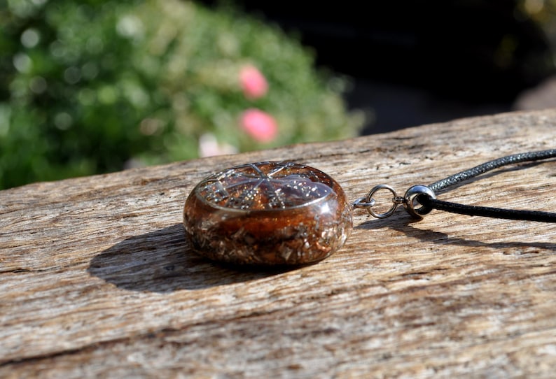 Op de afbeelding: Een ronde hanger met een heldere harskoepel gevuld met koperkleurige metaalvlokken en een zilverkleurige stervormige inleg. De hanger is bevestigd aan een dun zwart koord met een zilverkleurige metalen ring en een kleine cilindrische kraal.