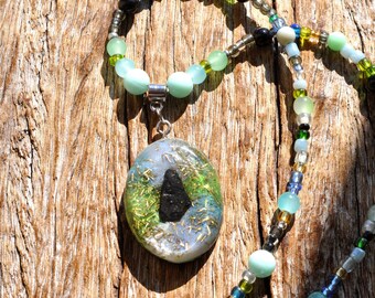 May include: A necklace featuring an oval pendant with a clear resin base containing green and gold metallic flecks and a central black stone fragment. The chain is composed of various small beads in shades of light blue, green, yellow, and black.