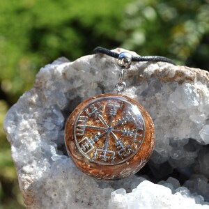 Op de afbeelding: Een ronde hanger met een zilverkleurig Vegvisir-symbool ingebed in een doorschijnende, amberkleurige harsbasis. De hanger is bevestigd aan een zwart koord met een zilverkleurig metalen oogje en toont een gedetailleerd kompasontwerp.