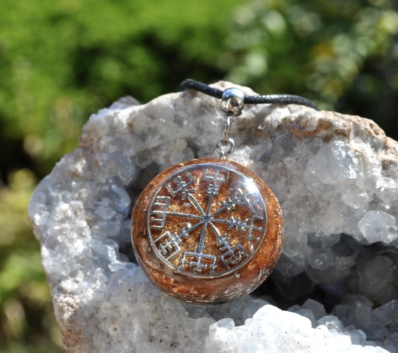 Op de afbeelding: Een ronde hanger met een zilverkleurig Vegvisir-symbool ingebed in een doorschijnende, amberkleurige harsbasis. De hanger is bevestigd aan een zwart koord met een zilverkleurig metalen oogje en toont een gedetailleerd kompasontwerp.