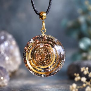 May include: A circular pendant featuring a gold-toned concentric ring design embedded within a clear resin disc. The pendant is suspended from a black cord by a gold-toned metal bail, showcasing metallic flakes and reflective elements.