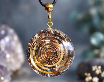 May include: A circular pendant featuring a gold-toned concentric ring design embedded within a clear resin disc. The pendant is suspended from a black cord by a gold-toned metal bail, showcasing metallic flakes and reflective elements.