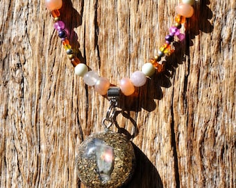 May include: A necklace featuring a spherical pendant filled with gold-toned metallic flakes and a central iridescent stone fragment. The pendant hangs from a strand of multi-colored beads in shades of peach, purple, yellow, and translucent white.