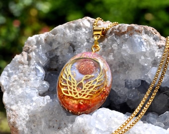 May include: An oval pendant necklace featuring a gold-toned phoenix emblem set against a background of orange and pink resin with embedded glitter. The pendant hangs from a gold-toned metal chain, showcasing a detailed bird with wings spread.