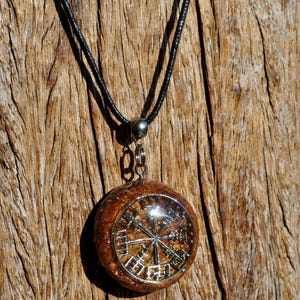 Op de afbeelding: Een ronde hangerketting met een zilverkleurig Vegvisir-symbool ingebed in een getextureerde, barnsteenkleurige harsbasis. De hanger is bevestigd aan een eenvoudig zwart koord met een zilverkleurig metalen oogje, wat een cirkelvormig middelpunt vormt.