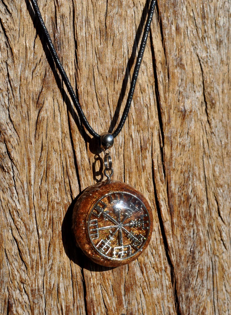 Op de afbeelding: Een ronde hangerketting met een zilverkleurig Vegvisir-symbool ingebed in een getextureerde, barnsteenkleurige harsbasis. De hanger is bevestigd aan een eenvoudig zwart koord met een zilverkleurig metalen oogje, wat een cirkelvormig middelpunt vormt.