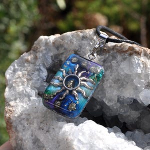 May include: A rectangular pendant with a silver sun design, featuring blue, green, and purple hues. The pendant is attached to a black cord. The pendant is set in a clear resin, with gold flecks.