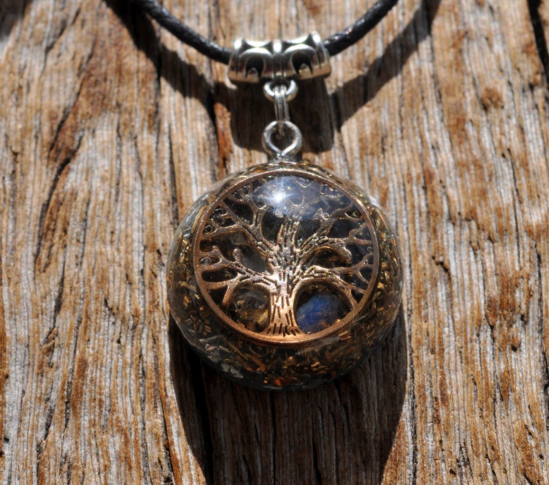 Puede incluir: Un collar con un colgante redondo de resina transparente que contiene un dise&ntilde;o de &aacute;rbol de la vida dorado y cristales azules y plateados. El collar est&aacute; en un cord&oacute;n negro.