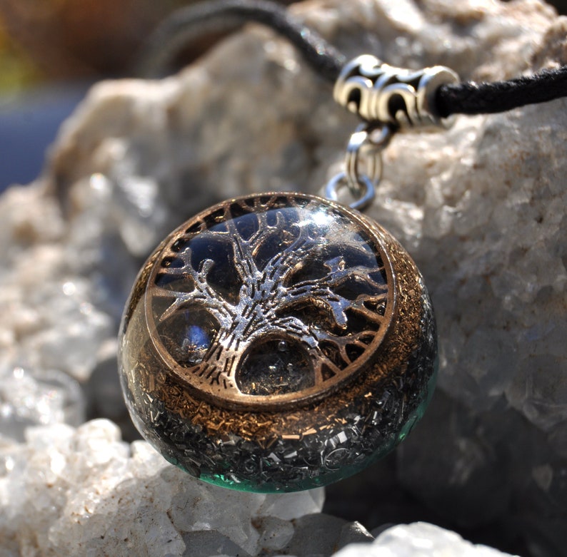 Puede incluir: Un colgante de &aacute;rbol de la vida de plata y oro con fondo negro, encapsulado en resina transparente y colocado en un cord&oacute;n negro. El colgante est&aacute; rodeado de peque&ntilde;as motas plateadas y doradas brillantes.