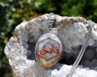 May include: An oval pendant necklace featuring a clear resin base embedded with gold-toned metal shavings and a central brown stone fragment. A gold-toned hexagonal frame sits atop the resin, suspended from a silver-toned metal chain.