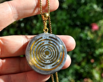 May include: A circular pendant featuring a gold-colored labyrinth design embedded in a translucent, light gray, speckled resin base. The pendant hangs from a gold-colored metal box chain, with a small gold-colored bail connecting the two pieces.