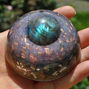 May include: A dome-shaped resin paperweight featuring a central polished labradorite gemstone with blue iridescence. The clear resin body is embedded with purple glitter, small copper metal shavings, and metallic flakes throughout the structure.