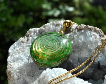 May include: A round green pendant featuring a gold-colored concentric circle design on its surface. The pendant is attached to a thin gold-colored metal chain and rests on a textured, crystalline, light-colored mineral base.