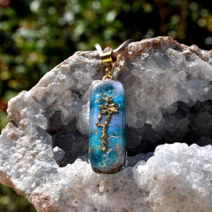 May include: A rectangular pendant with a gold-colored tree design encased in a clear resin. The resin has blue and teal hues, and the pendant hangs from a brown cord. The background is a white and gray crystal formation.