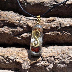 May include: A rectangular pendant necklace with a gold infinity symbol encased in clear resin. The pendant contains small red and gold-colored stones and is suspended from a black cord.