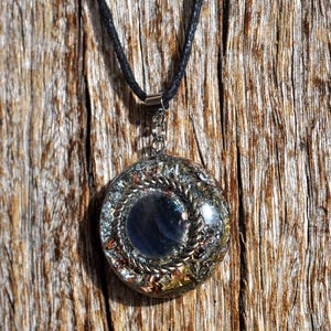 May include: A circular pendant necklace featuring a dark blue stone at its core, encircled by silver and copper-toned metal detailing. It is suspended from a black cord. The backdrop is a textured wooden surface.