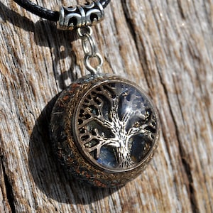 Puede incluir: Un collar con un colgante de plata redondo, de resina transparente, que contiene un dise&ntilde;o de &aacute;rbol de la vida en plata. La esfera est&aacute; rodeada por un borde de metal de color cobre y tiene un acabado brillante e iridiscente.
