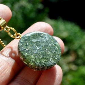 Op de afbeelding: Een ronde hanger met een doorschijnende olijfgroene harsbasis ingelegd met glinsterende zilverkleurige metaalvlokken en fijne glitterdeeltjes, bevestigd aan een goudkleurige metalen ring en een bijpassende goudkleurige schakelketting.