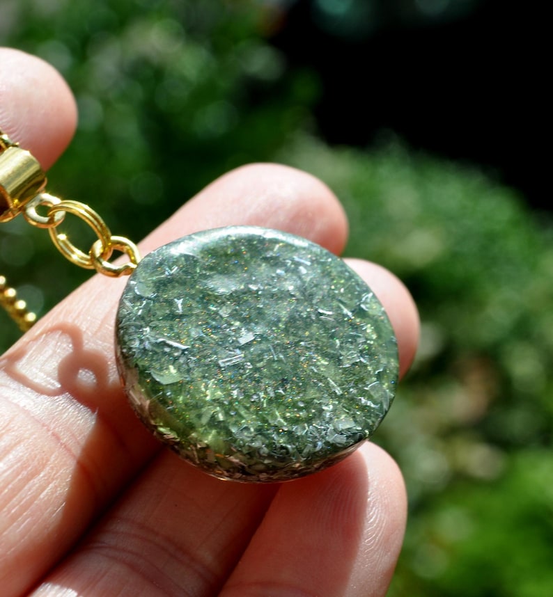 Op de afbeelding: Een ronde hanger met een doorschijnende olijfgroene harsbasis ingelegd met glinsterende zilverkleurige metaalvlokken en fijne glitterdeeltjes, bevestigd aan een goudkleurige metalen ring en een bijpassende goudkleurige schakelketting.