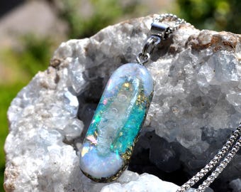 May include: An oval-shaped pendant featuring a mix of iridescent blue, green, and white flecks with gold-colored accents, suspended from a silver-toned metal chain. The pendant has a smooth, glossy finish and a rounded, pill-like silhouette.