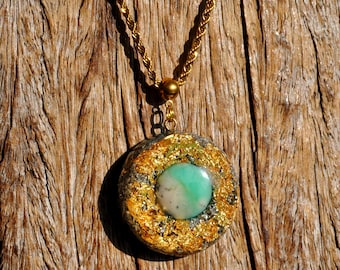 May include: A circular pendant necklace featuring a central round turquoise and white stone, surrounded by a textured layer of gold leaf flakes and dark grey metallic particles, suspended from a gold-toned twisted rope chain.
