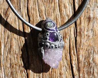 May include: A pendant necklace featuring a rough, light purple druzy crystal base set in a textured, silver-toned metal frame. The frame is adorned with a small, round, iridescent stone and a smooth, oval, deep purple cabochon stone.