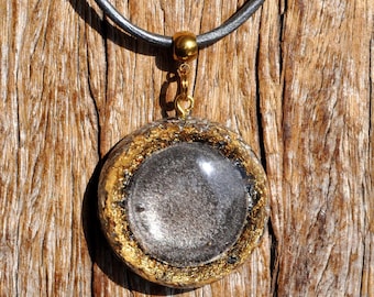 May include: A circular pendant featuring a dark, shimmering gray center surrounded by a textured gold-colored border. The pendant is attached to a smooth, dark gray cord by a polished gold-toned metal bail and a small jump ring.