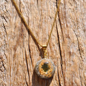 Op de afbeelding: Een goudkleurige boxketting met een ronde hanger. De hanger is gemaakt van heldere hars met daarin metallic goudvlokken en een donkergroen steenfragment in het midden, opgehangen aan een goudkleurig metalen oogje en een ketting.
