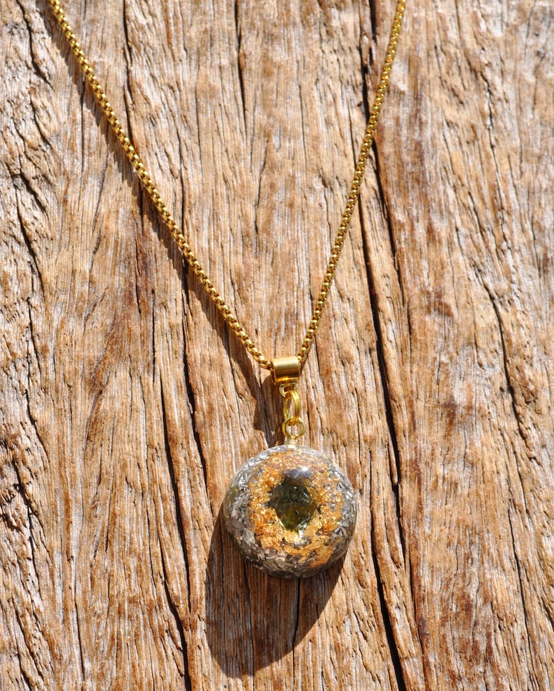 Op de afbeelding: Een goudkleurige boxketting met een ronde hanger. De hanger is gemaakt van heldere hars met daarin metallic goudvlokken en een donkergroen steenfragment in het midden, opgehangen aan een goudkleurig metalen oogje en een ketting.