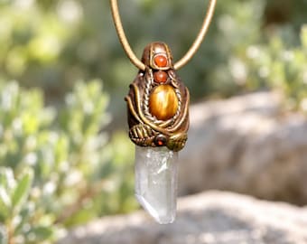 May include: A pendant necklace featuring a clear, pointed quartz crystal suspended from a tan cord. The setting is crafted from bronze-toned metal with sculpted leaf details, a central oval tiger's eye stone, and two small round orange beads.