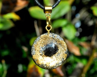 May include: A circular pendant featuring a central black stone surrounded by gold leaf flakes and silver metallic fragments, suspended from a black cord by a gold-toned metal bail and jump ring, creating a textured, multi-material appearance.