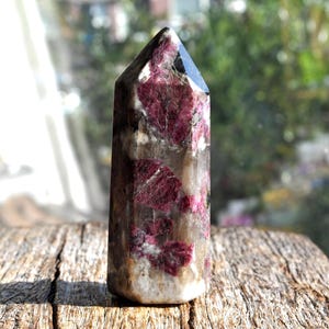 May include: A polished hexagonal crystal tower featuring a translucent quartz base with prominent, deep magenta-colored inclusions throughout. The stone displays a pointed top and a smooth, multifaceted surface that catches the light.