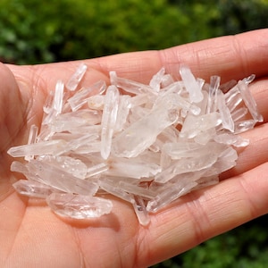 May include: A pile of clear quartz crystal points, also known as rock crystals, in a hand. The points are various sizes and shapes, with some being long and thin, while others are shorter and thicker.