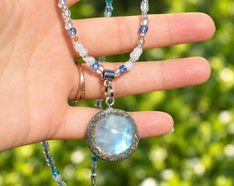 May include: A necklace featuring a round, iridescent moonstone-like pendant set in a textured, dark metallic frame. The chain is composed of small, multi-colored glass beads in shades of white, light blue, and dark blue, with a silver-tone clasp.